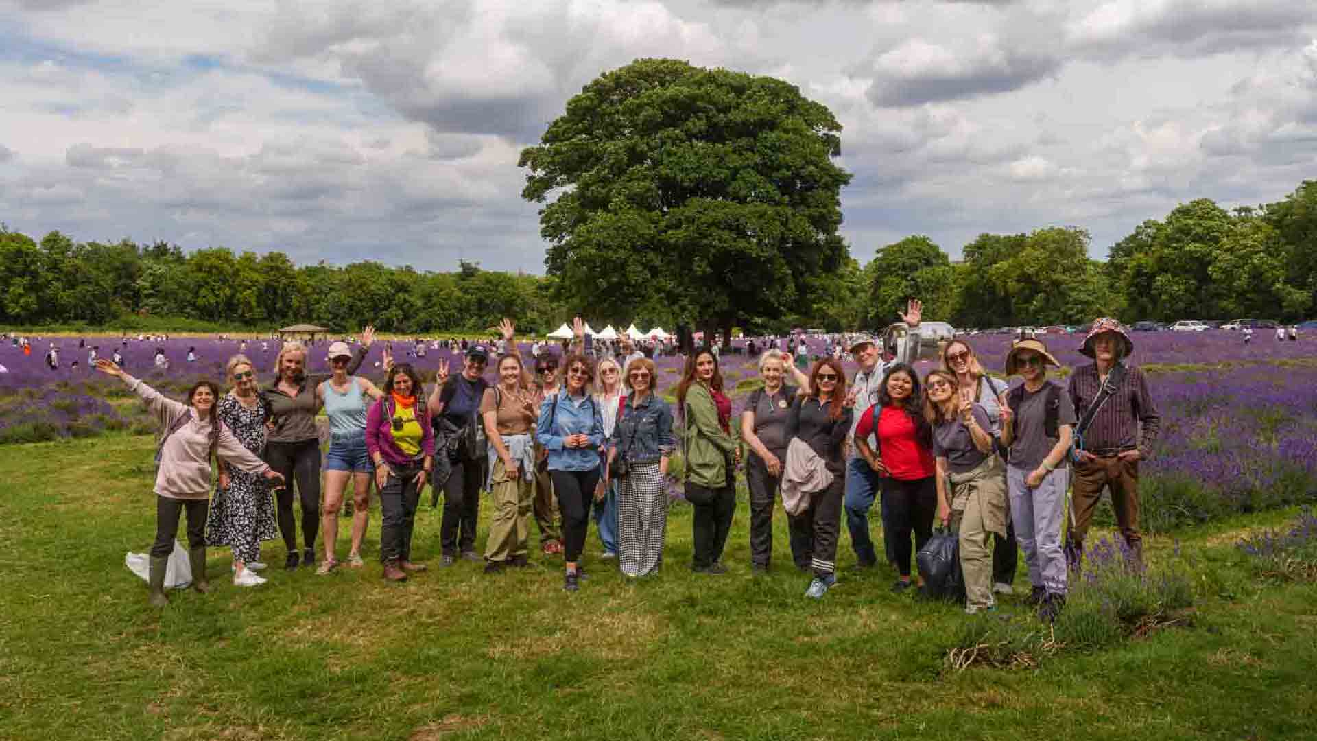 A group of hikers smiling and posing together at Mayfield Lavender Farm, surrounded by vibrant purple lavender fields and lush green trees under a cloudy sky.