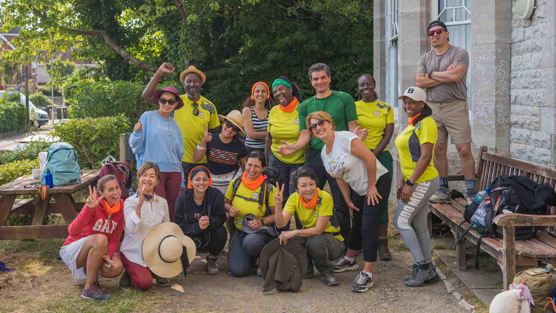 A diverse group of hikers from the Hiking in London club posing together outdoors. Some members are wearing bright yellow hiking shirts with the club logo, while others are in casual attire. The group is smiling, making peace signs