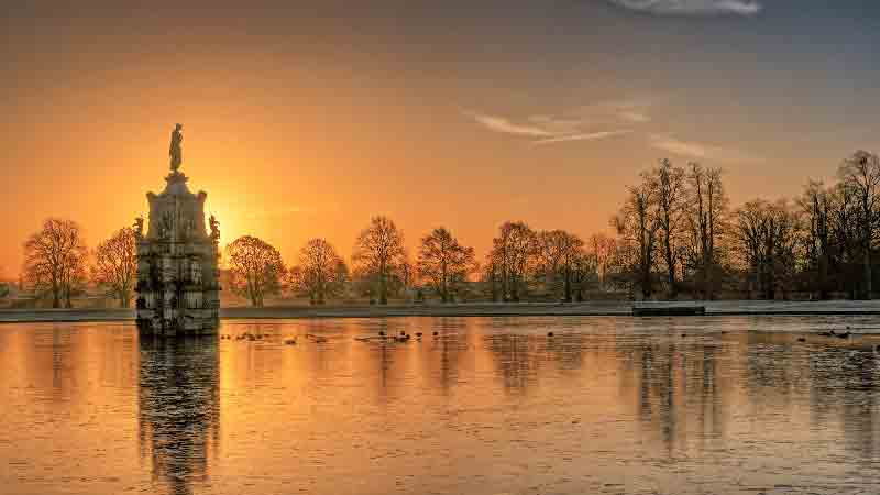 Sunset view of the Diana Fountain at the center of Chestnut Avenue in Bushy Park, reflecting on a calm pond surrounded by bare trees