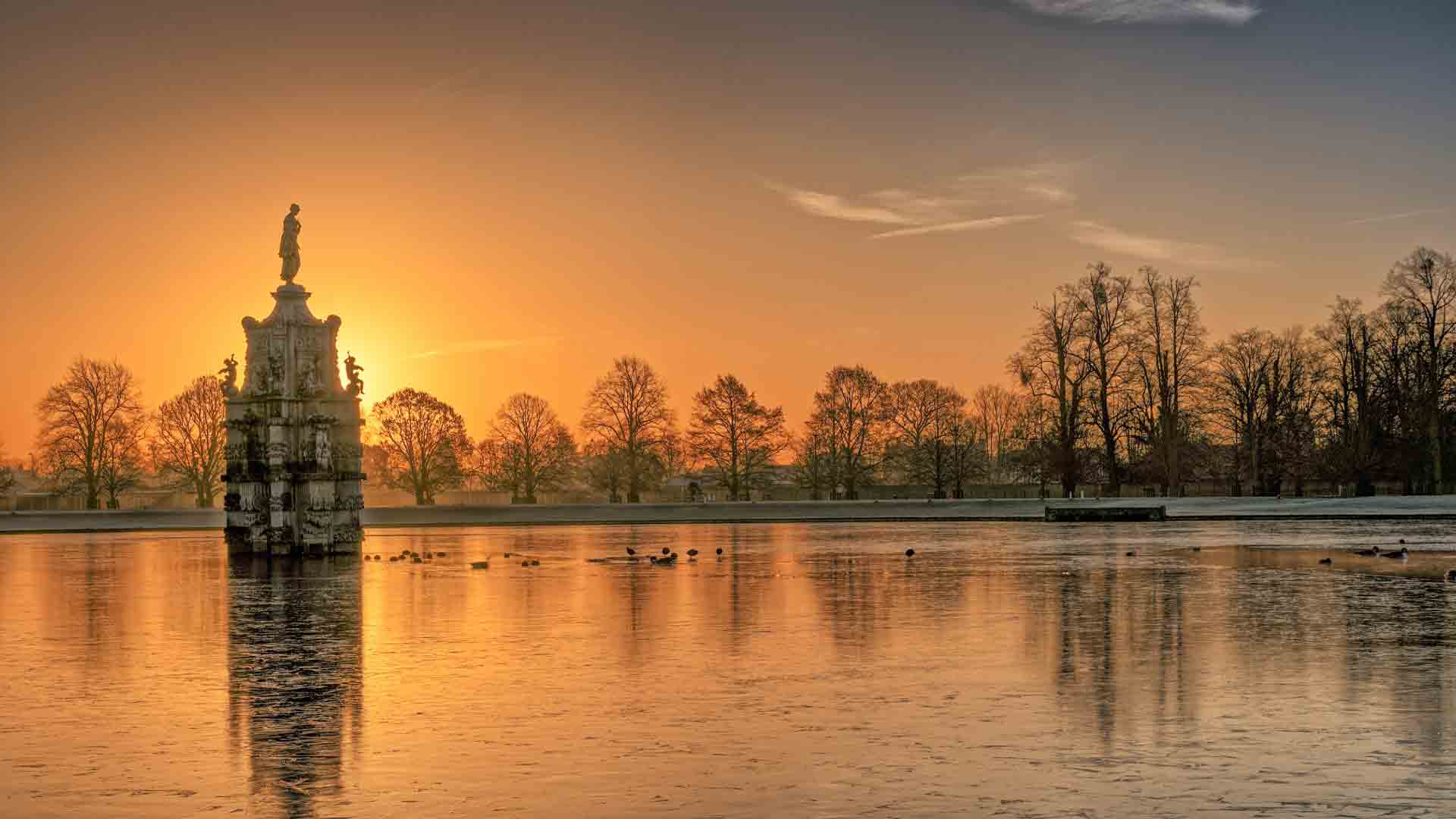 Sunset view of the Diana Fountain at the center of Chestnut Avenue in Bushy Park, reflecting on a calm pond surrounded by bare trees