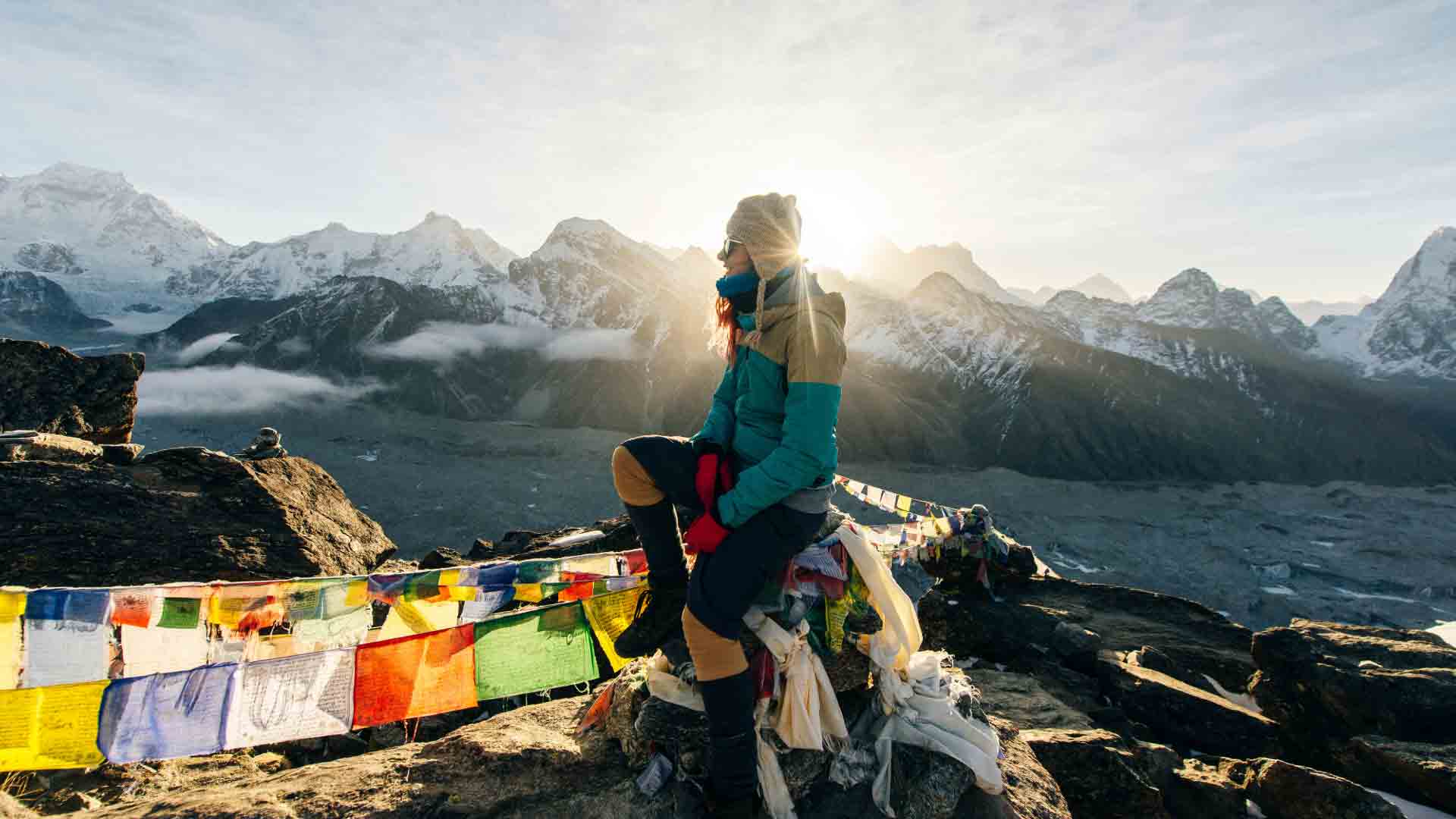 Trekker seated on rocky terrain at sunrise, surrounded by colorful prayer flags and Himalayan peaks