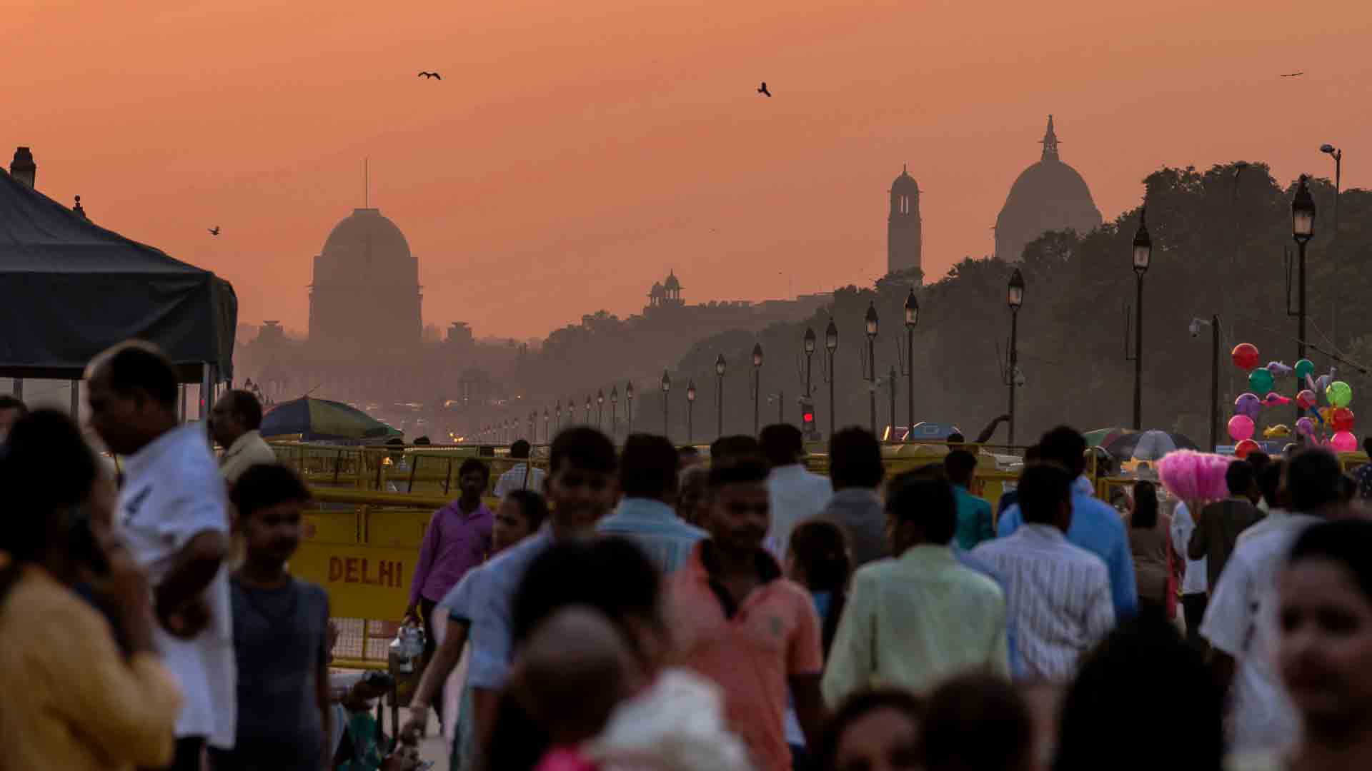 Crowded street in New Delhi at sunset with silhouettes of iconic buildings and lively street life.